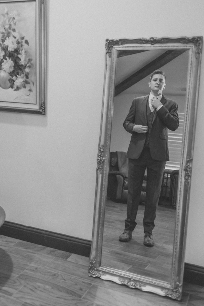 Groom fixing his tie in a tall standing mirror all in black and white as if frozen in time