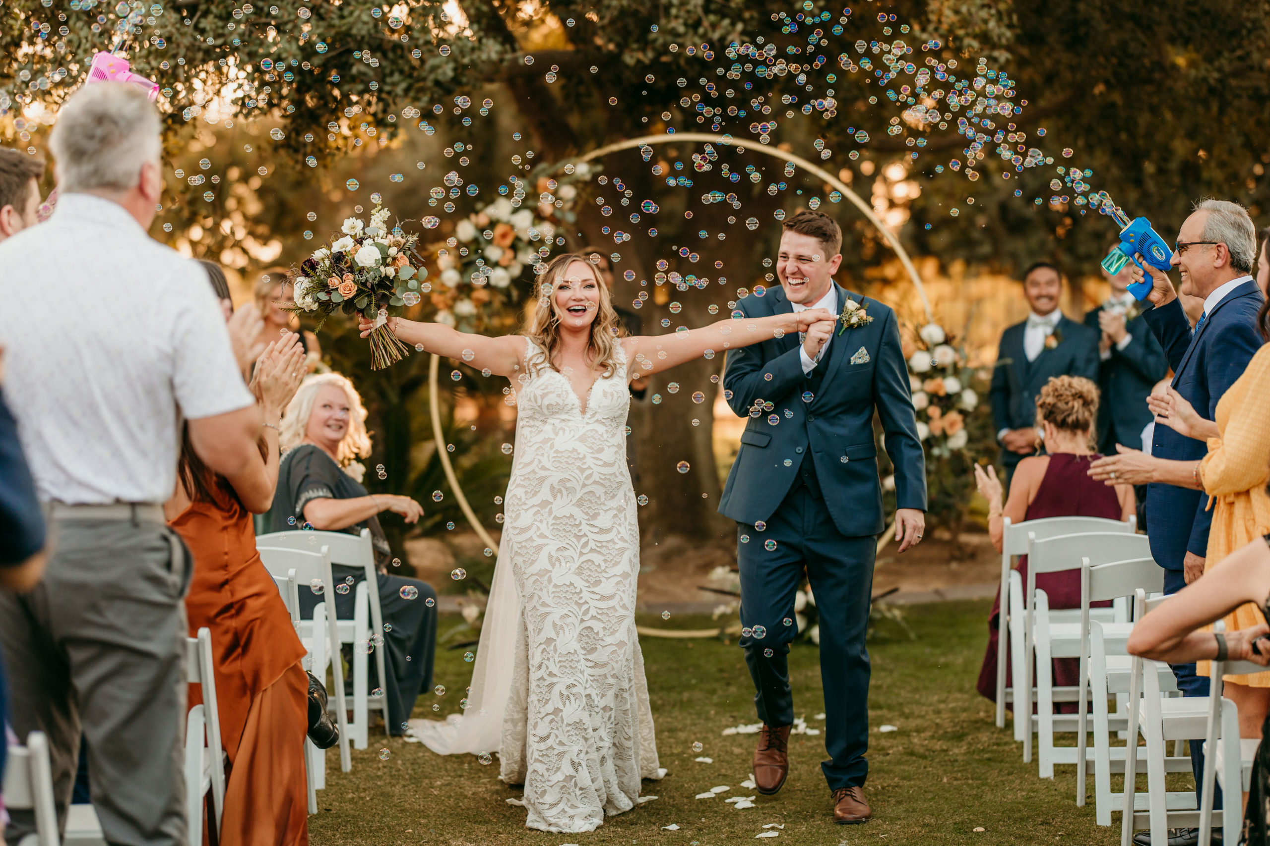 Newly married couple walking back up the aisle through a cloud of glittery bubbles. Bride is cheering with her arms open wide and groom is laughing a joyous laugh.